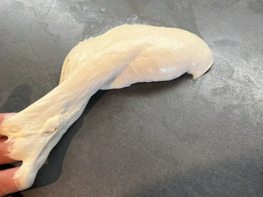 Ein Stück heller, klebriger Pizzateig wird von einer Hand gehalten und dehnt sich auf einer dunklen Arbeitsfläche aus.