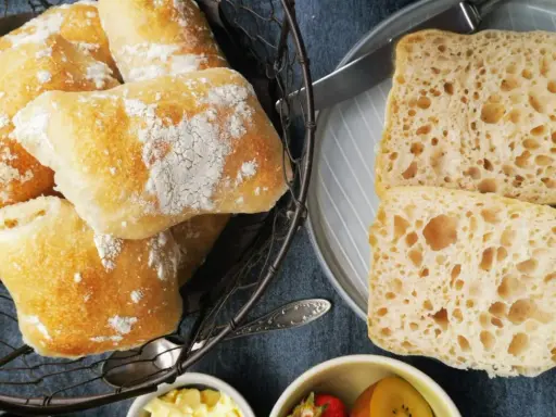 Frisch gebackene Ciabatta-Brötchen, bestäubt mit Mehl, liegen in einem Korb. Daneben zwei aufgeschnittene Scheiben mit luftiger Krume – perfekt zum ciabatta selber backen.