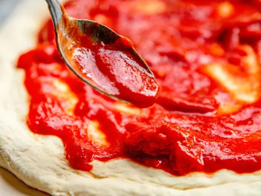 Eine Löffelspitze verteilt die leuchtend rote Tomatensauce auf dem Teig für eine authentische Pizza Napoletana.