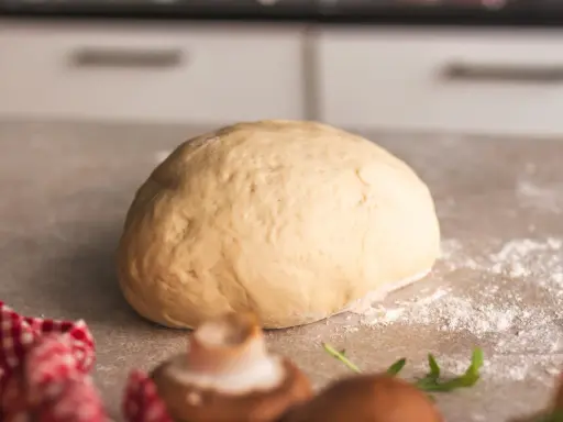 Frischer, goldgelber Pizzateig liegt bereit zum Formen. Ein paar Pilze und Rucola deuten auf eine leckere Pizza hin.