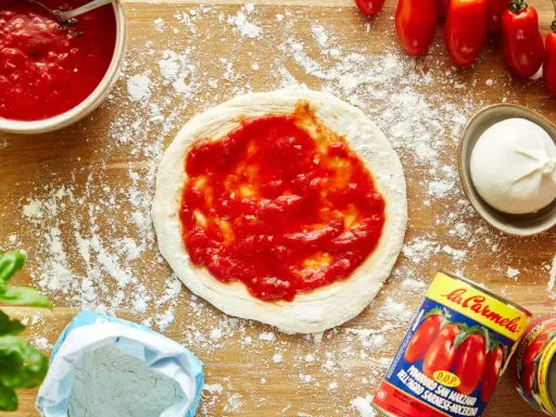 Pizza-Zutaten auf Holzbrett: Teig mit Tomatensauce, Mozzarella, frische Tomaten, Basilikum, Mehl und eine Dose San Marzano Tomaten. Die Frage "wieviel Tomatensauce pro Person" wird hier visuell beantwortet.