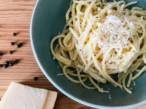 Ein Teller cremiges Cacio e Pepe, bestreut mit frisch geriebenem Pecorino und schwarzem Pfeffer. Daneben liegen Pfefferkörner und ein Stück Pecorino.