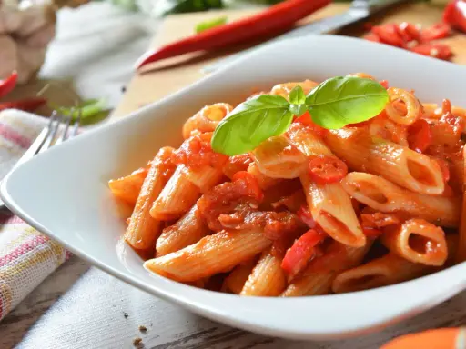 Eine Schüssel Penne Arrabiata, garniert mit Basilikum, mit scharfen Chilischoten und Knoblauch im Hintergrund.