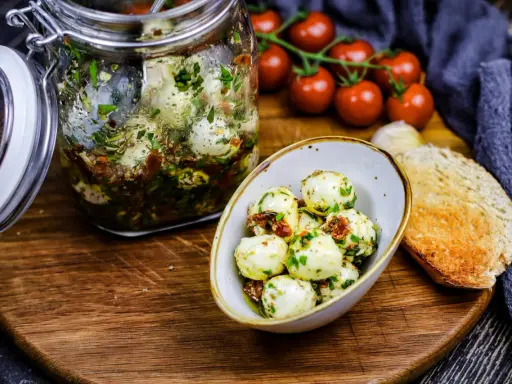 Marinierte Mozzarellakugeln in einem Glas, bereit für köstliche Mozzarella Rezepte. Dazu gibt es frische Tomaten und geröstetes Brot.