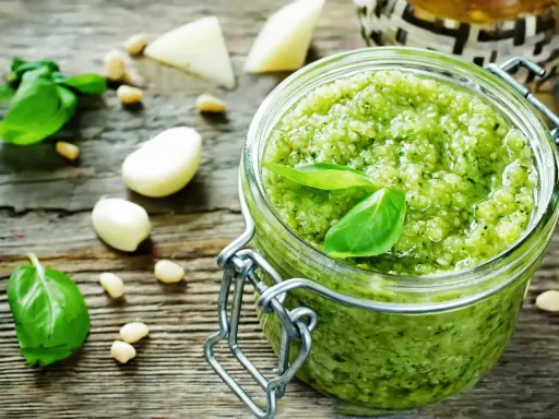 Frisches Pesto-Rezept im Glas, umgeben von Basilikum, Pinienkernen, Knoblauch und Parmesan auf rustikalem Holz.