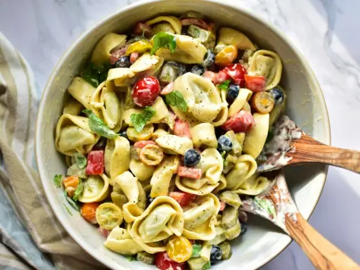 Ein cremiger Tortellini Salat mit Kirschtomaten, Oliven und Kräutern, serviert in einer Schüssel mit Holzspateln.