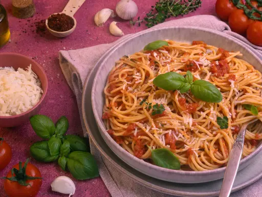 Ein Teller Spaghetti Napoli mit Tomatensauce, Basilikum und Parmesan. Daneben frische Tomaten, Knoblauch und Kräuter.