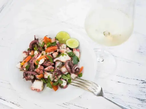 Frischer Insalata di polpo mit Tomaten, Petersilie und Limettenspalten auf einem Teller, daneben ein Glas Weißwein.