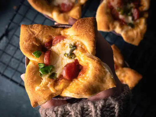 Hand hält einen herzhaften Pizza-Muffin mit geschmolzenem Käse, Wurst und grünen Paprika.