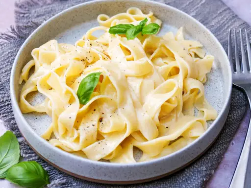 Cremige Pasta Alfredo mit frischem Basilikum und schwarzem Pfeffer, serviert auf einem grauen Tischtuch.