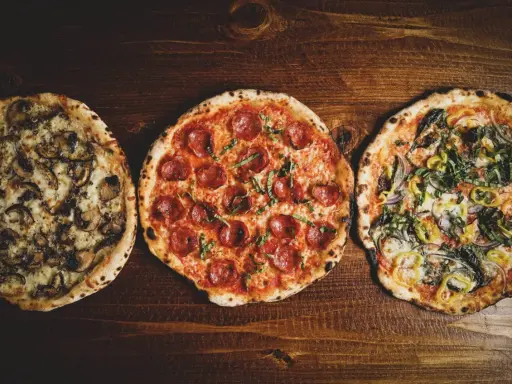 Drei leckere Pizza-Sorten: eine mit Pilzen, eine mit Salami und eine mit Gemüse, liegen nebeneinander auf einem Holztisch.