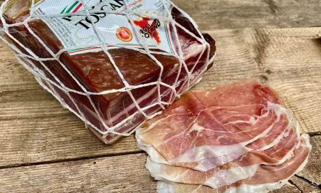 Ein Block **Prosciutto Toscano** im Netz und daneben dünn geschnittene Scheiben.