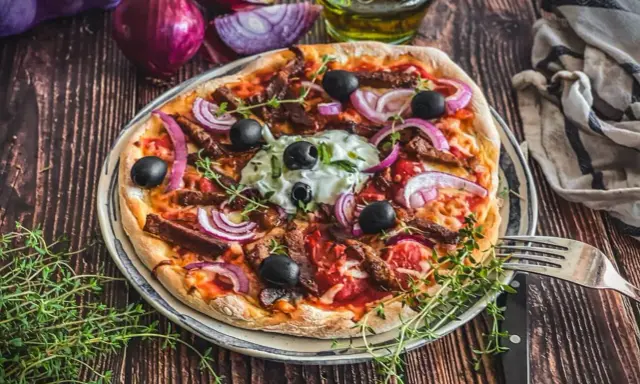 Herzhafte Gyros Pizza mit Zwiebeln, Oliven und Kräutern, serviert auf einem Teller mit Gabel und Messer.