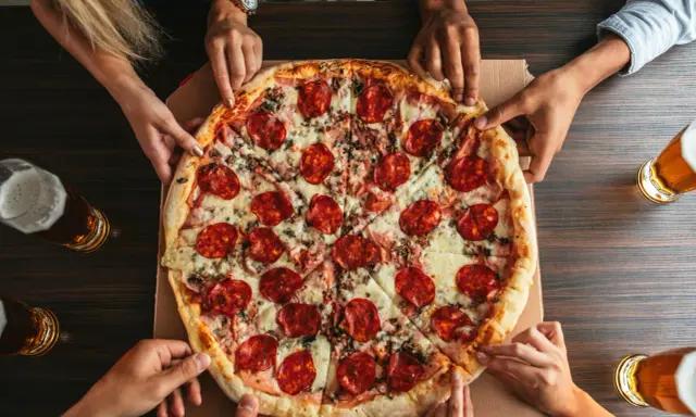Hände greifen nach einer großen Salamipizza, die auf einem Tisch liegt. Jeder bekommt seinen fairen Anteil an pizzateig pro person.