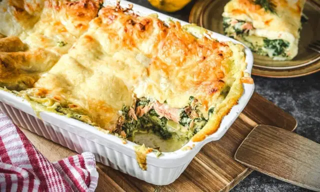 Herzhafte Lachs Spinat Lasagne, goldbraun überbacken, dampft in einer weißen Auflaufform. Ein Stück auf einem Teller daneben.