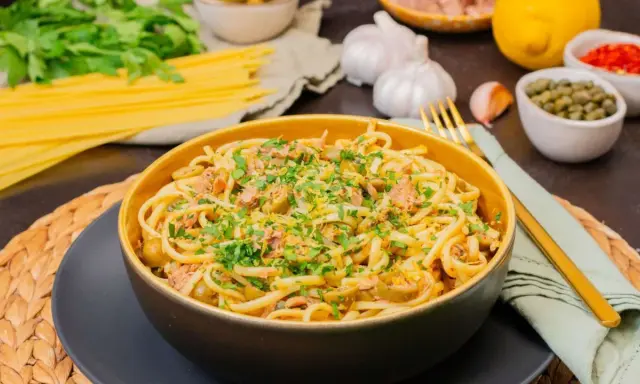 Eine Schüssel mit köstlicher Thunfisch Pasta, garniert mit Petersilie und Zitronenschale. Daneben liegen Spaghetti, Knoblauch und Kapern.