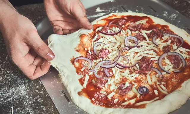 Hände schieben einen rohen Pizzaboden mit Tomatensauce, Käse, Salami und Zwiebeln auf einen Pizzaschieber. Der pizzateig klebt am schieber.