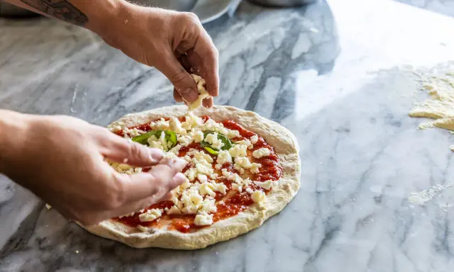 Hände belegen Pizza mit zerbröckeltem Käse, einer köstlichen fior di latte Alternative, auf Marmortisch.