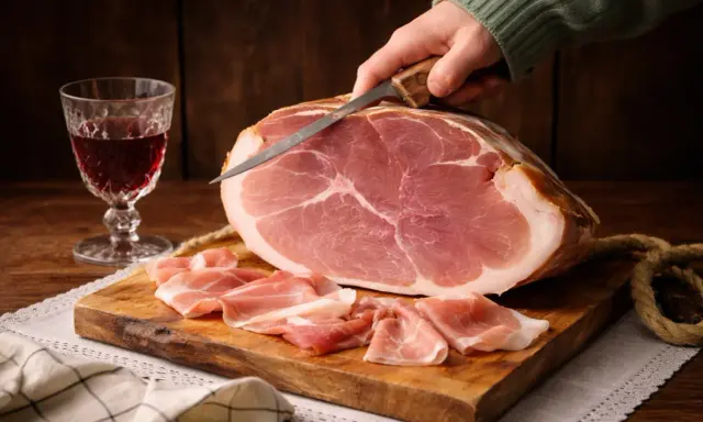 Ein Messer schneidet hauchdünne Scheiben von einem ganzen Schinken, **prosciutto crudo**, auf einem Holzbrett. Daneben ein Glas Rotwein.