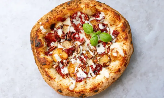 Pizza mit karamellisierten Zwiebeln, Speck, Pekannüssen und einem Hauch von Parmesan. Ein grünes Basilikumblatt krönt das Kunstwerk.