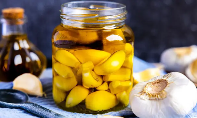 Ein Glas mit eingelegtem Knoblauch, typisch italienisch, gefüllt mit goldgelbem Öl. Daneben frische Knoblauchzehen und eine Ölflasche.