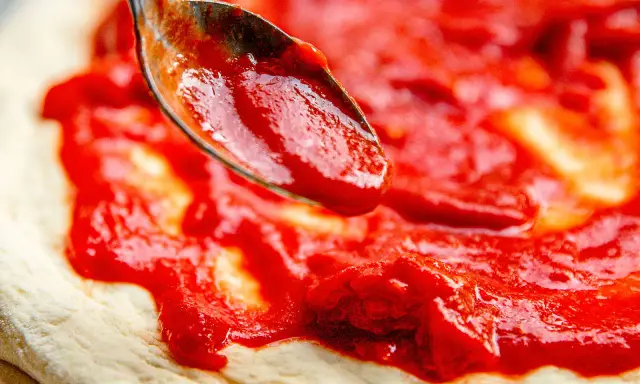 Eine Löffelspitze verteilt die leuchtend rote Tomatensauce auf dem Teig für eine authentische Pizza Napoletana.