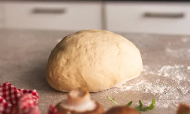 Frischer, goldgelber Pizzateig liegt bereit zum Formen. Ein paar Pilze und Rucola deuten auf eine leckere Pizza hin.
