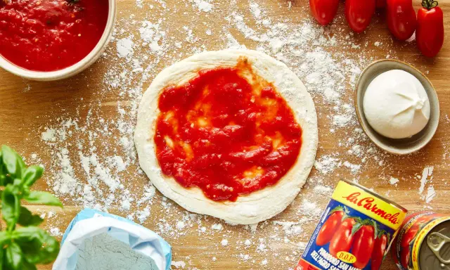 Pizza-Zutaten auf Holzbrett: Teig mit Tomatensauce, Mozzarella, frische Tomaten, Basilikum, Mehl und eine Dose San Marzano Tomaten. Die Frage "wieviel Tomatensauce pro Person" wird hier visuell beantwortet.