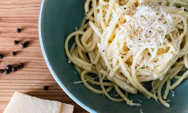 Ein Teller cremiges Cacio e Pepe, bestreut mit frisch geriebenem Pecorino und schwarzem Pfeffer. Daneben liegen Pfefferkörner und ein Stück Pecorino.