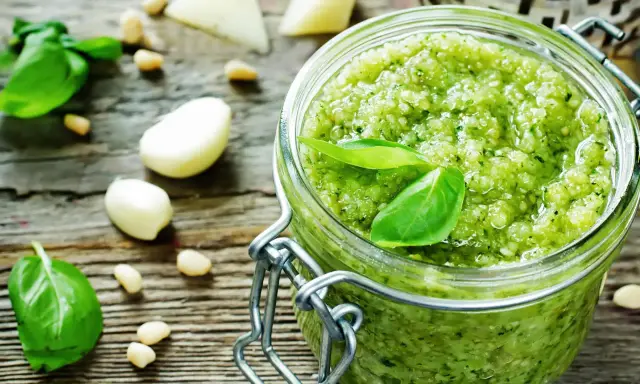 Frisches Pesto-Rezept im Glas, umgeben von Basilikum, Pinienkernen, Knoblauch und Parmesan auf rustikalem Holz.