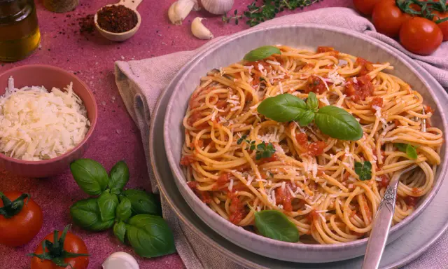 Ein Teller Spaghetti Napoli mit Tomatensauce, Basilikum und Parmesan. Daneben frische Tomaten, Knoblauch und Kräuter.