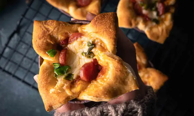Hand hält einen herzhaften Pizza-Muffin mit geschmolzenem Käse, Wurst und grünen Paprika.
