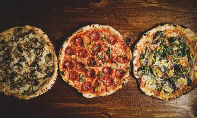 Drei leckere Pizza-Sorten: eine mit Pilzen, eine mit Salami und eine mit Gemüse, liegen nebeneinander auf einem Holztisch.