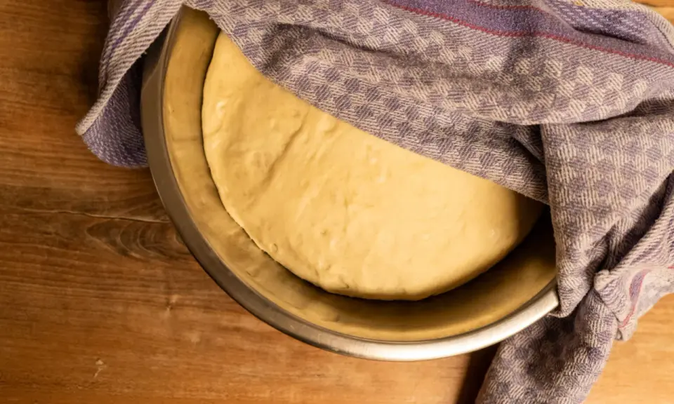 Frischer, aufgegangener Pizzateig ruht in einer Schüssel, bedeckt mit einem gemusterten Tuch. Perfekt für Ihr Pizzateig Rezept.