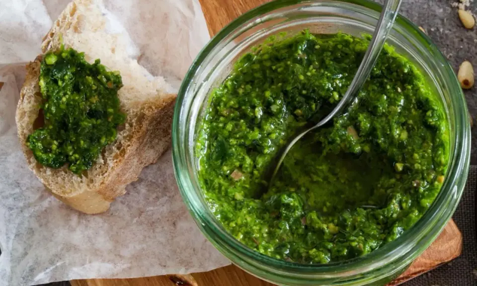 Frisches schnittlauch pesto in einem Glas, mit einem Löffel darin. Ein Stück Brot mit dem grünen Pesto darauf liegt daneben.