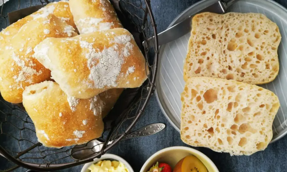 Frisch gebackene Ciabatta-Brötchen, bestäubt mit Mehl, liegen in einem Korb. Daneben zwei aufgeschnittene Scheiben mit luftiger Krume – perfekt zum ciabatta selber backen.