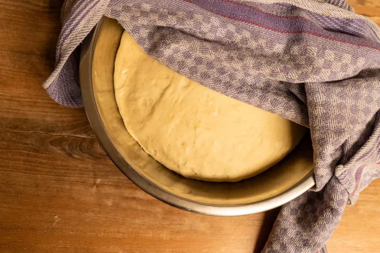 Frischer, aufgegangener Pizzateig ruht in einer Schüssel, bedeckt mit einem gemusterten Tuch. Perfekt für Ihr Pizzateig Rezept.