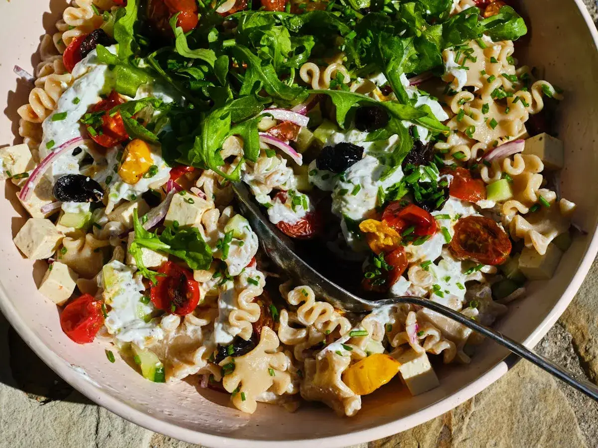 Ein bunter mediterraner Nudelsalat mit viel frischem Gemüse, Oliven und cremiger Soße, garniert mit Rucola und Schnittlauch.