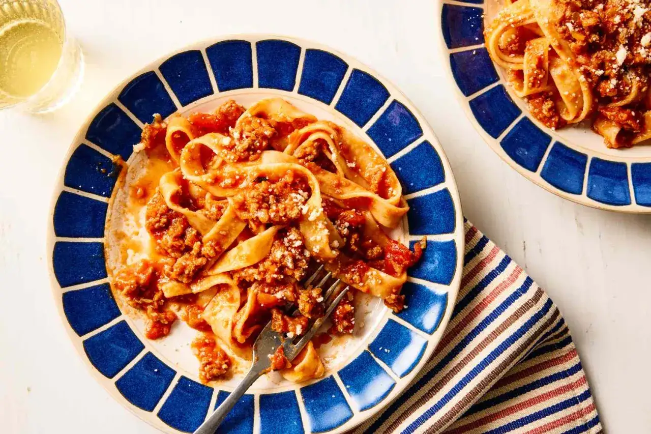 Teller mit breiten Bandnudeln und einer herzhaften, schnellen Bolognese Sauce, bestreut mit Parmesan. Ein Glas Wei&szlig;wein steht daneben.