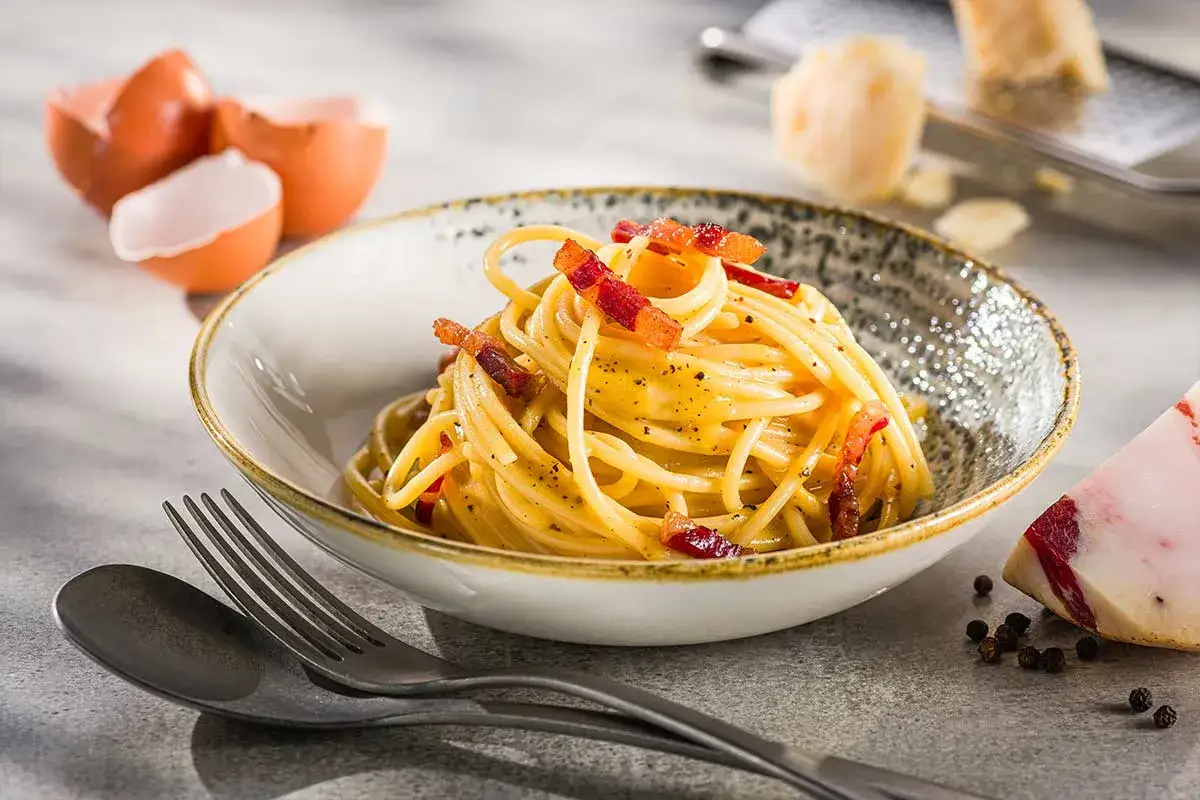 Eine Sch&uuml;ssel cremiger Spaghetti Carbonara, garniert mit knusprigem Speck und frisch gemahlenem Pfeffer. Daneben liegen Eierschalen und ein St&uuml;ck Pecorino.