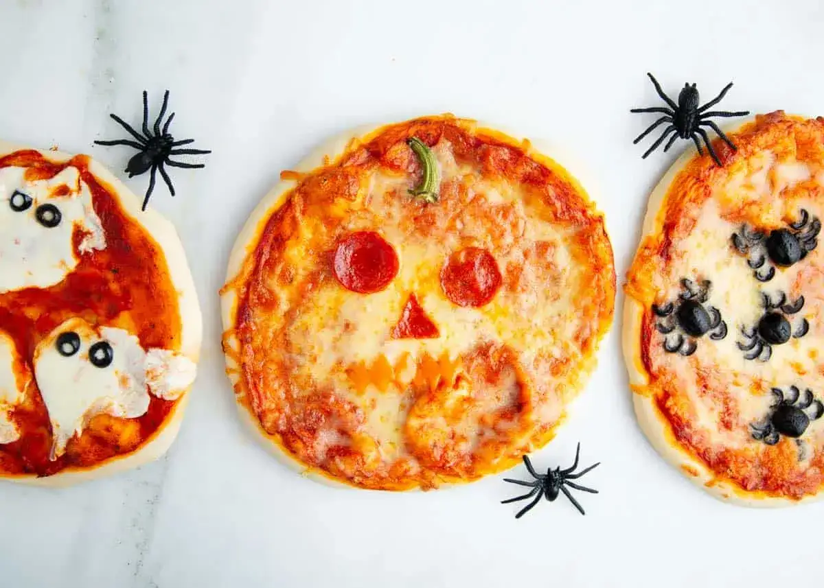Drei lustige Halloween-Pizzen: eine als K&uuml;rbisgesicht, eine als Geist und eine mit Spinnen aus Oliven. Perfekt f&uuml;r eine gruselige Feier!