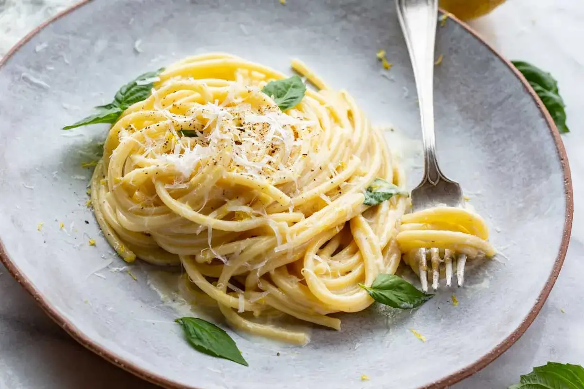 Ein Teller cremiger Spaghetti al Limone, garniert mit Parmesan, Basilikum und Zitronenschale. Eine Gabel nimmt eine Portion auf.