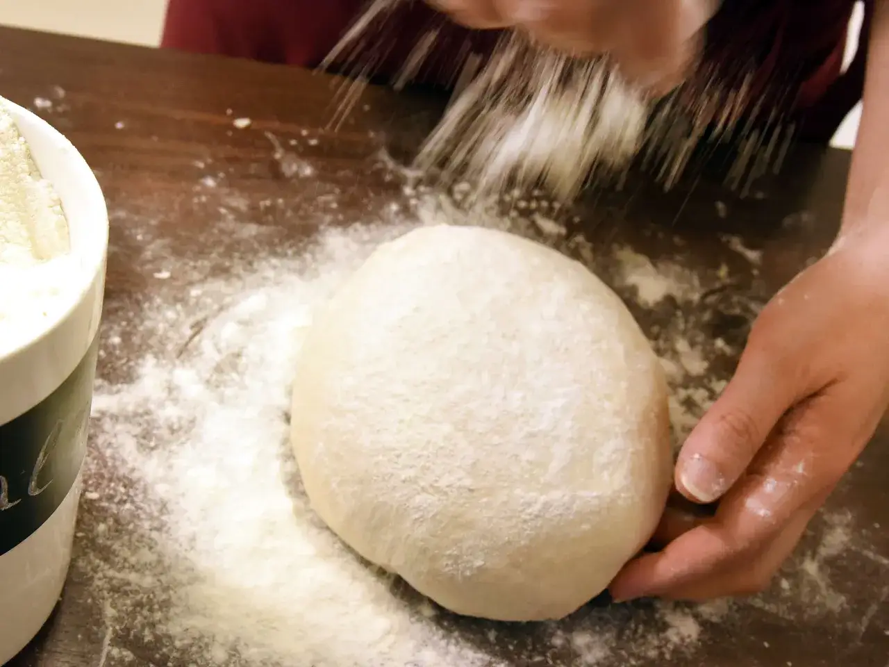 Eine Hand streut Mehl über einen Teigballen. Nebenan steht eine Schüssel mit self-raising flour deutsch.