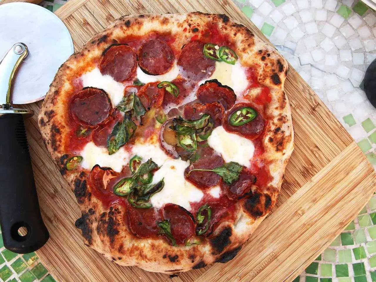 Eine scharfe Salami Pizza mit Jalape&ntilde;os und Mozzarella auf einem Holzbrett, daneben ein Pizzaschneider.