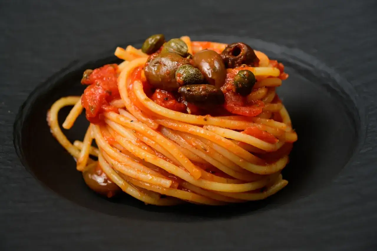 Ein kunstvoll aufget&uuml;rmter Teller Spaghetti Puttanesca, garniert mit Oliven und Kapern, auf schwarzem Teller.