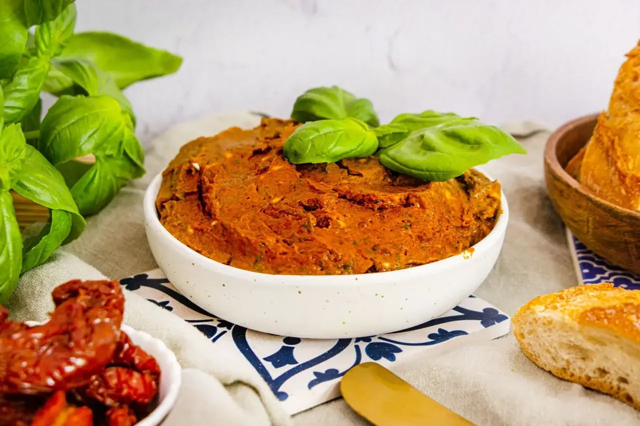 Herzhaftes Tomatenbutter Rezept in einer Sch&uuml;ssel, garniert mit Basilikum. Daneben liegen getrocknete Tomaten und Baguettescheiben.