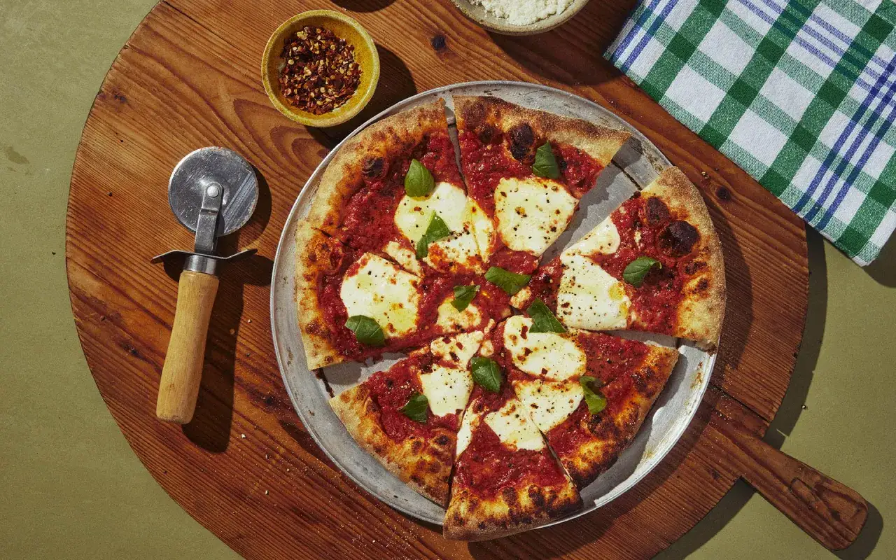 Eine Margherita-Pizza mit reichlich San Marzano Pizza Sauce, Mozzarella und Basilikum auf einem Holzbrett, daneben ein Pizzaschneider und Chiliflocken.