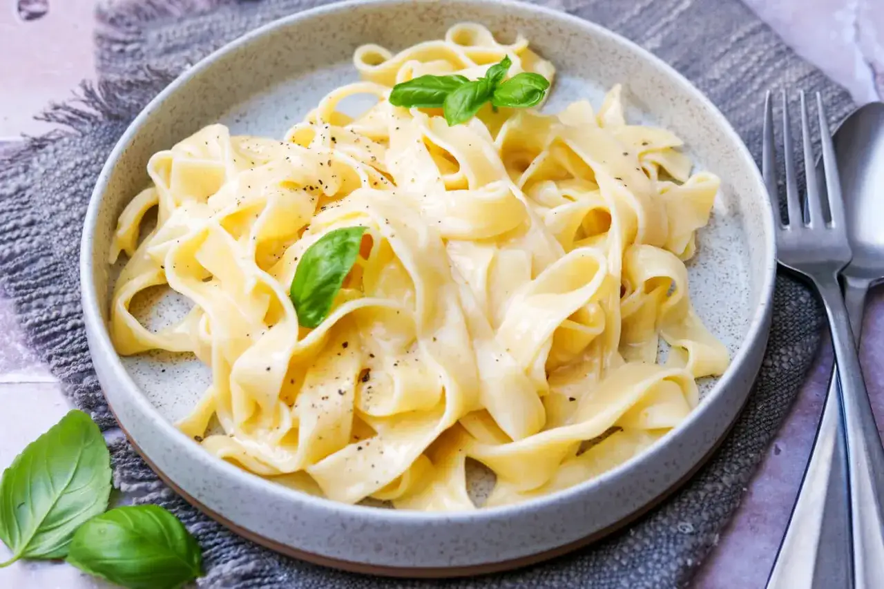 Cremiges Pasta Alfredo mit frischem Basilikum und schwarzem Pfeffer, serviert in einer grauen Schüssel auf einem rustikalen Tuch.