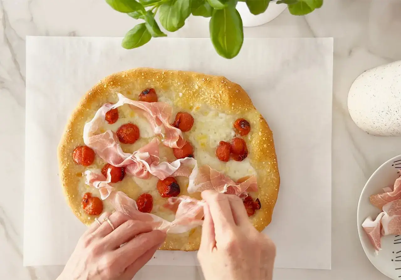 Hände legen dünne Scheiben prosciutto schinken auf eine Focaccia mit Kirschtomaten und Käse.
