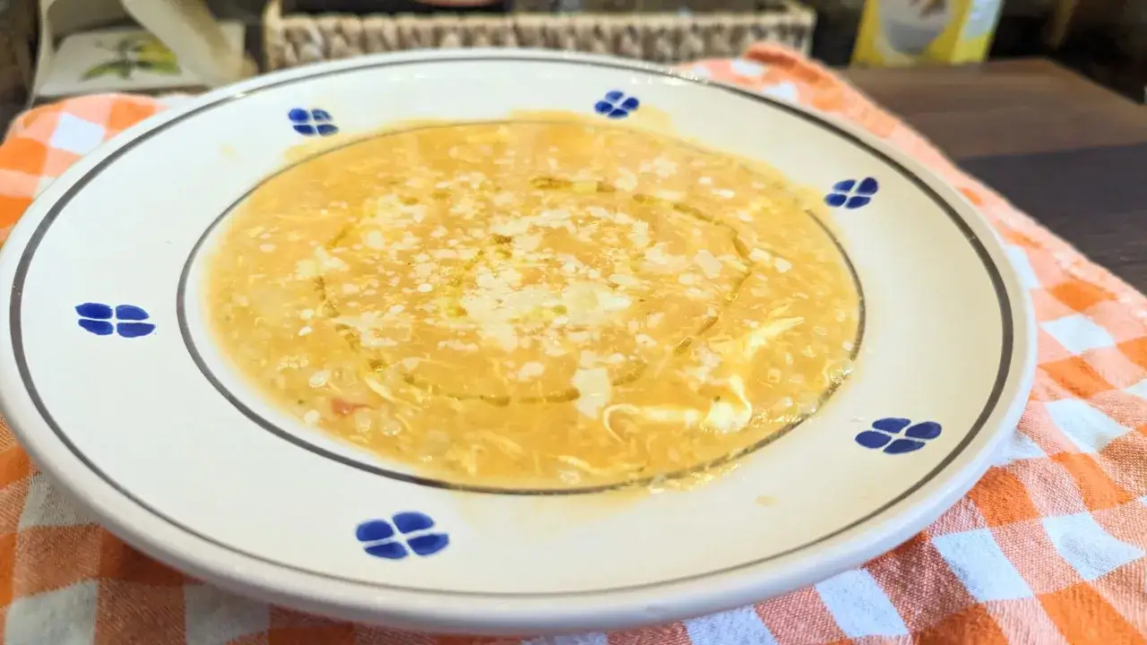Eine dampfende Sch&uuml;ssel Pastina Suppe, garniert mit Parmesan und einem Hauch Oliven&ouml;l, auf einem karierten Tuch.